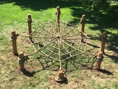 an outdoor structure made out of sticks and ropes in the grass with trees behind it