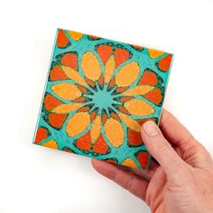 a hand holding up a small square tile with an orange and blue flower on it