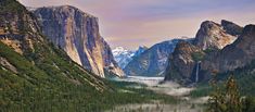 the valley is surrounded by tall mountains and snow capped peaks, with mist in the foreground