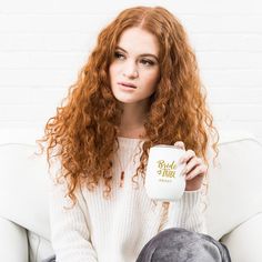 a woman sitting on a white couch holding a coffee mug in her right hand and looking at the camera