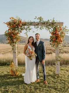 a bride and groom pose for a photo under an arch decorated with orange flowers at their wedding