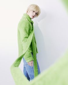 a woman standing in front of a white wall wearing a green sweater and jeans with her hands on her hips