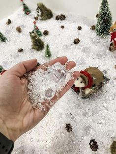 a hand is holding an ice cube with small toy animals in the snow behind it