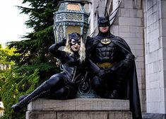 two people dressed up as batman and catwoman sitting on a stone bench in front of a building