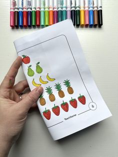 a hand holding a paper with fruit and vegetables on it next to crayons