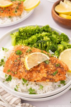 two plates with fish, rice and broccoli next to lemon wedges on the table