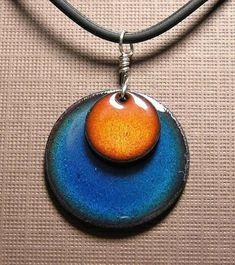 an orange and blue pendant hangs from a leather cord on a tan surface with a black cord