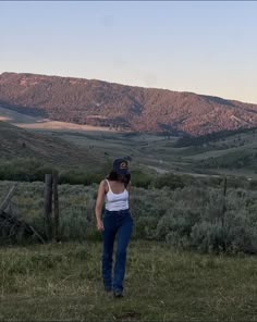 a woman standing on top of a lush green field next to a mountain covered in trees