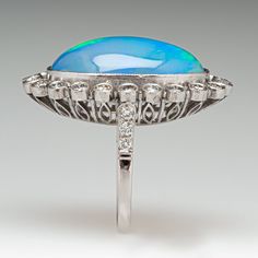 This gorgeous ring is centered with one (1) oval cabochon cut natural crystal opal and set into a bezel setting. The opal is bordered with twenty-two (22), bezel set, round brilliant cut diamonds, The top face of the shank is accented with three (3), bead set, round brilliant cut diamonds on each side. The gemstones are bordered with milgrain edging. The ring measures 29.1mm at the top, rises 10.6mm above the finger, tapering to 1.8mm wide and 1.1mm thick at the base of the shank. This ring is currently a size 6. Fine Jewelry Oval Cabochon Opal Ring, Oval Cabochon Opal Ring Fine Jewelry, Fine Jewelry Oval Opal Ring With Polished Finish, Oval Cabochon Opal Ring In Fine Jewelry, Formal Oval Cabochon Opal Ring, Oval Opal Ring With Polished Finish In Fine Jewelry, Oval Opal Ring With Polished Finish, Classic Opal Ring With Diamond Accents, Exquisite Multi-stone Oval Opal Ring