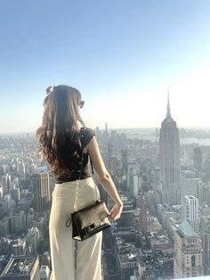 a woman standing on top of a tall building looking at the city below her and holding a purse