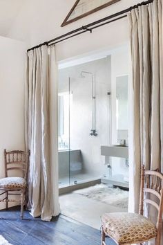 a bathroom with a shower, toilet and chair next to a window that has drapes on it