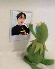 a green stuffed animal sitting next to a white wall with an image of kermie the frog on it