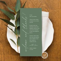 a green wedding program on a white plate with eucalyptus leaves in the center and an olive branch next to it