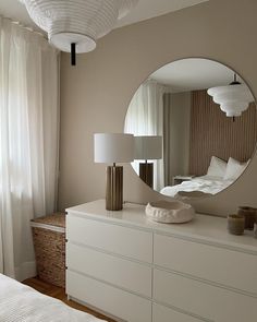 a white dresser topped with a round mirror next to a window covered in curtains and pillows