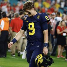 a football player walking on the field with his helmet in hand and people behind him