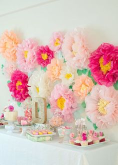 a white table topped with lots of pink and yellow flowers next to a wall covered in paper flowers