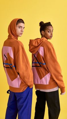 two people standing next to each other in front of a yellow background wearing orange and pink hoodies