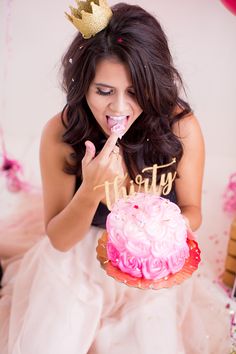 a woman in a white dress eating a pink frosted cake with a gold tiara on her head