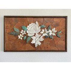 a wall hanging with white flowers and green leaves on brown stone tiles in a wood frame