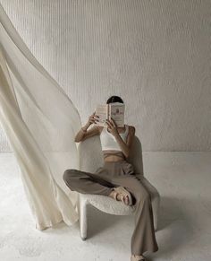 a woman sitting in a chair reading a book while wearing brown pants and a white tank top