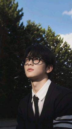 a young man wearing glasses and a black suit is standing in front of some trees