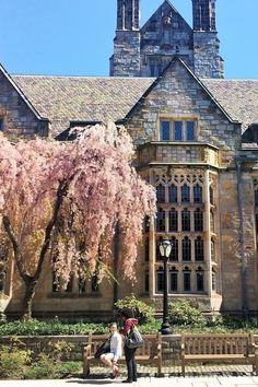 two people sitting on a bench in front of a large building with trees and flowers