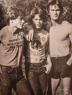 three young men standing next to each other in front of some trees and grass with one man pointing at the camera