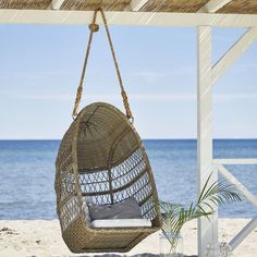 a rattan hanging chair on the beach next to the ocean with a pillow in it