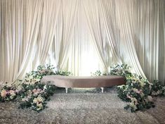 a couch sitting in front of a window covered with white drapes and floral arrangements