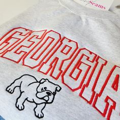 Experience the perfect blend of style and school spirit with our Georgia Bulldog Crewneck Sweatshirt or T-shirt! Available in three colors, this sweatshirt is designed to keep you cozy and comfortable, whether you're cheering in the stands or just showing off your Bulldog pride around town. At the heart of its design is the iconic "Georgia" embroidery, boldly presented across the center chest. The word is intricately stitched in a vibrant red, capturing the essence of the University of Georgia's Collegiate Cotton Sweatshirt With Screen Print, Varsity Cotton Sweatshirt With Screen Print, Varsity Tops With Embroidered Graphics For Campus, Varsity Tops With Embroidered Logo For Campus, Varsity Style Embroidered Tops For Campus, College Cotton Sweatshirt With Screen Print, Collegiate Cotton Sweatshirt With University Logo, College Sweatshirt With Embroidered Logo, College Sweatshirt With Embroidered Logo For School Spirit