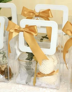 three clear boxes with gold ribbons and decorations in them on a tablecloth covered table