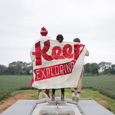 three people standing on the back of a truck holding a red and white sign that says keep exploring