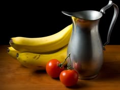 two bananas and three tomatoes sit on a table next to a silver pitcher with a metal lid