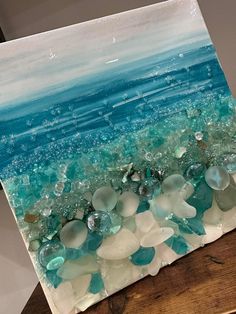 a painting with sea glass on the bottom and blue water in the middle, sitting on top of a wooden table