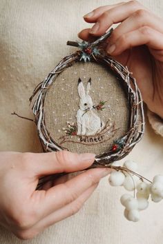 two hands holding a cross stitch ornament with a white rabbit on it and berries in the foreground