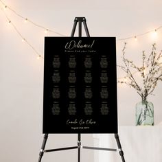 a black and white wedding seating chart on a easel with lights in the background