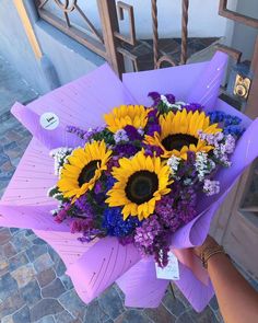 a bouquet of sunflowers and purple flowers is held by a woman's hand