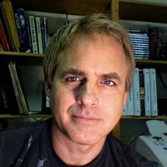 a man with blonde hair and blue eyes wearing a black shirt in front of a bookshelf