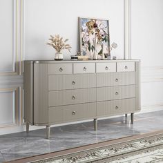 an ornate dresser with flowers on top in a white walled room, next to a painting and rug