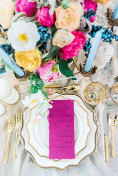 the table is set with pink and yellow flowers, silverware, and gold place settings