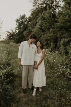 a man and woman standing next to each other in the grass with trees behind them