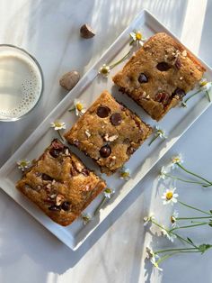 three squares of cake on a white plate next to a glass of milk and flowers