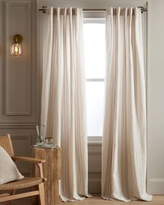 a white curtain hanging in front of a window next to a wooden chair and lamp