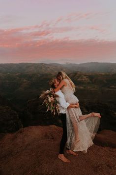 a bride and groom hugging on top of a mountain at sunset with the sun setting behind them