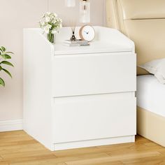 a white night stand with flowers and a clock on it next to a small bed