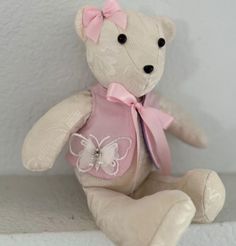 a white teddy bear with a pink shirt and butterfly on it's chest sitting against a wall