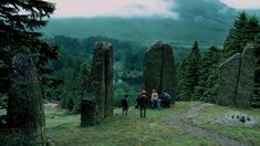 some people are standing in the grass near large rocks and trees with mountains in the background