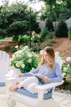 a woman sitting on a white chair reading a book in front of flowers and bushes