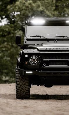 the front end of a black land rover with its lights on, parked in dirt area next to trees