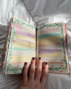 a woman's hand holding an open book on top of a white sheet covered bed